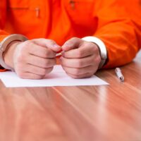 Close-up of a person in handcuffs wearing an orange jumpsuit, sitting at a table with paper and pen.