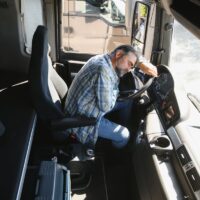 Person in plaid shirt sitting inside a truck cabin resting on the steering wheel.