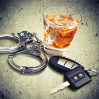 A glass of whiskey with ice, handcuffs, and a car key on a textured surface, symbolizing the dangers of drinking and driving.